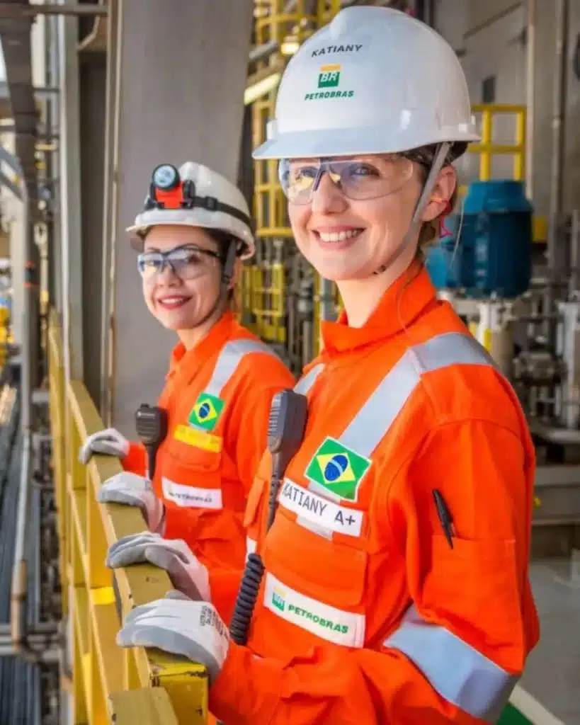 Petrobras oferece 60 vagas em curso gratuito para atuação no setor offshore: Duas mulheres, engenheiras ou técnicas, vestindo macacões laranja de segurança, capacetes e óculos de proteção, sorriem em uma plataforma de petróleo. Uma delas tem o nome "Katiany" no capacete e crachá, com o logotipo da Petrobras visível. A imagem destaca a presença feminina e a segurança no setor offshore de energia.
