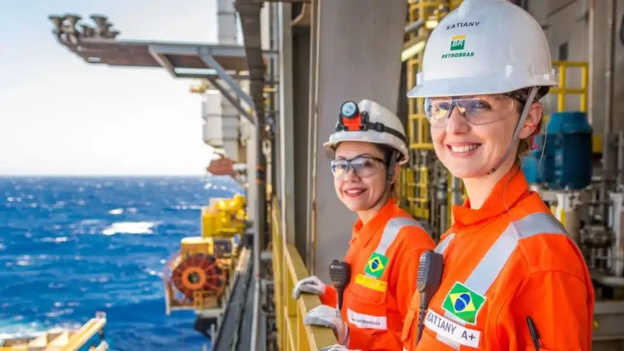 Petrobras oferece 60 vagas em curso gratuito para atuação no setor offshore 33 Duas mulheres em equipamentos de proteção individual (EPIs), incluindo macacões laranja e capacetes brancos, estão em uma estrutura offshore, com o oceano azul visível ao fundo. Uma delas, identificada como "Katiany", sorri para a câmera. A cena ilustra a atuação de profissionais femininas na indústria de petróleo e gás em alto mar.